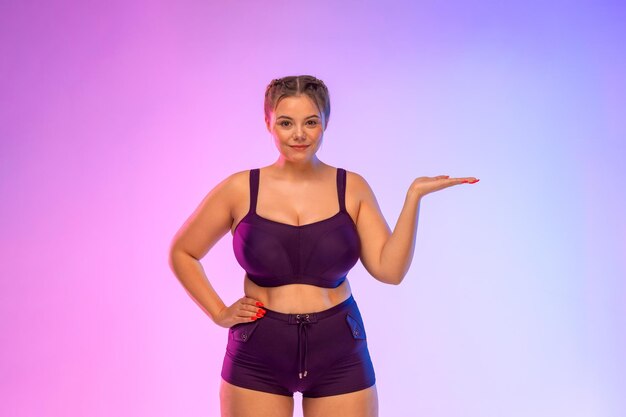 Body-positive woman standing on pink background, pointing at copy space.