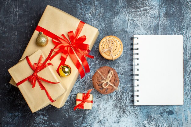 Wrapped gift boxes with red ribbons placed beside cookies and a blank notebook on a textured surface.