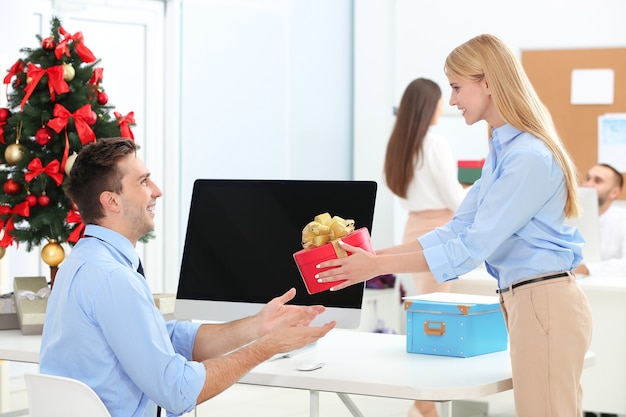 Office colleague presenting a gift to a seated coworker at a desk during a festive workplace setting.