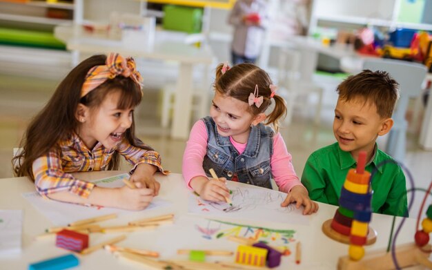 Three young children drawing and smiling together at a classroom table.
